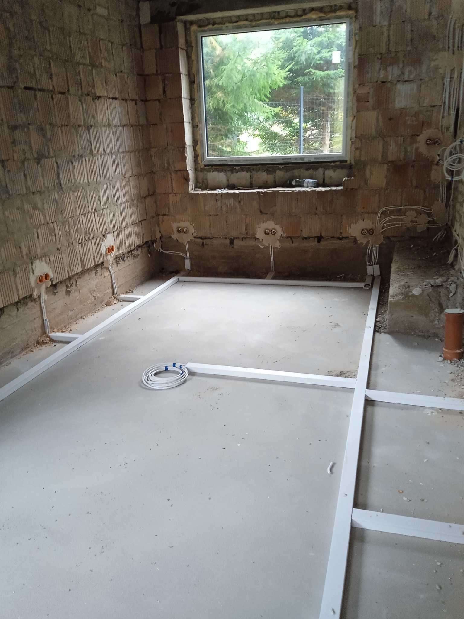 Wnętrze w trakcie remontu: surowe ściany z cegły, okno z widokiem na zieleń, przygotowane punkty elektryczne i ułożona konstrukcja pod podłogę.