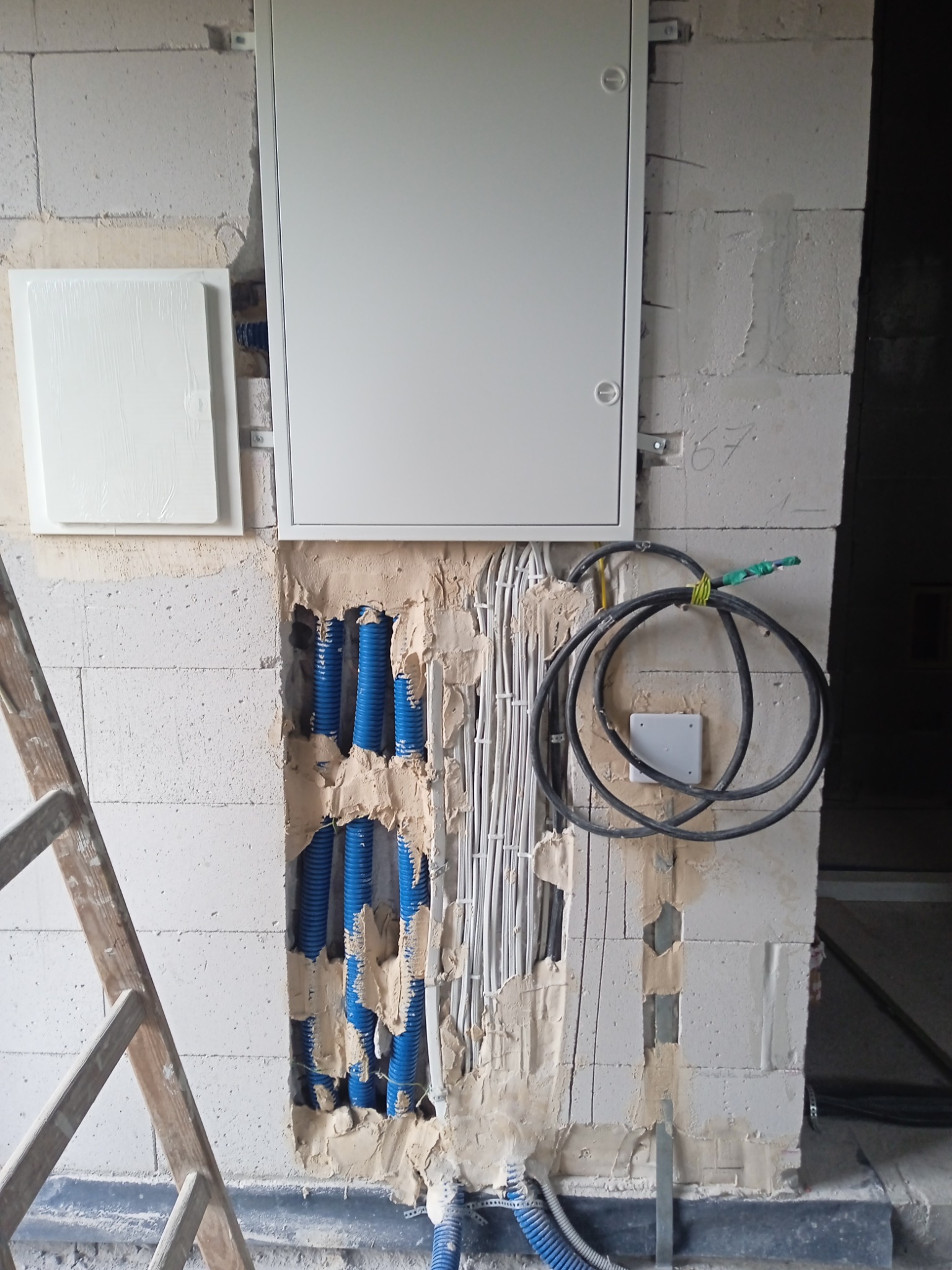 Instalacja elektryczna w ścianie z pustaków: biała skrzynka rozdzielcza, niebieskie rury karbowane, białe przewody. Widoczna drabina.