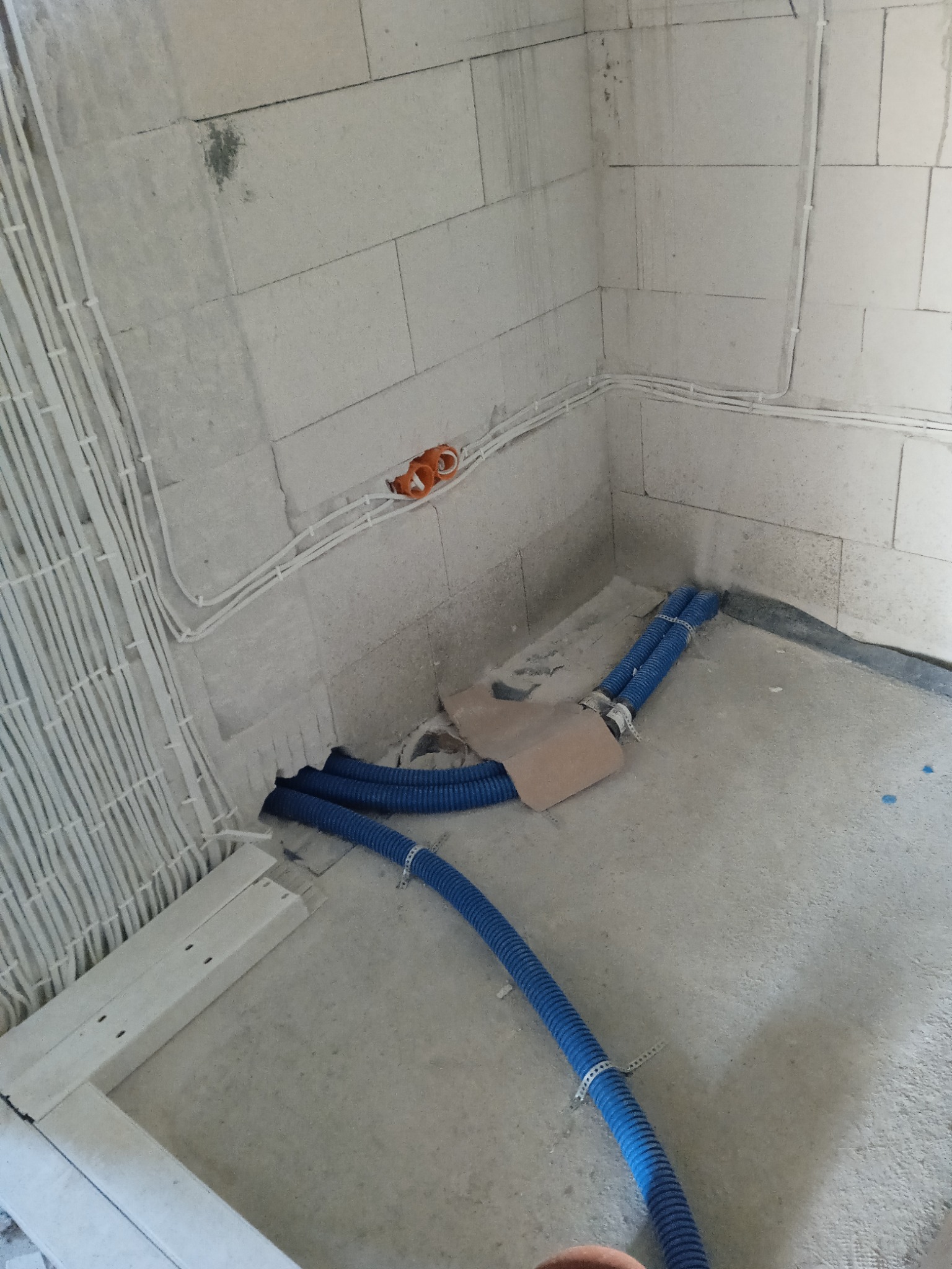 Surowy stan: kable elektryczne w białych rurkach na ścianie z pustaków, niebieskie rury w podłodze, pomarańczowe puszki elektryczne. Instalacja w trakcie prac.