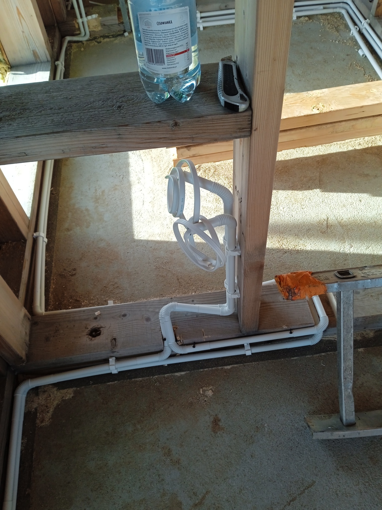 Białe rury instalacji wodnej zamocowane do drewnianej konstrukcji, widoczna butelka wody i nożyk. Instalacja w surowym stanie, przed wykończeniem.