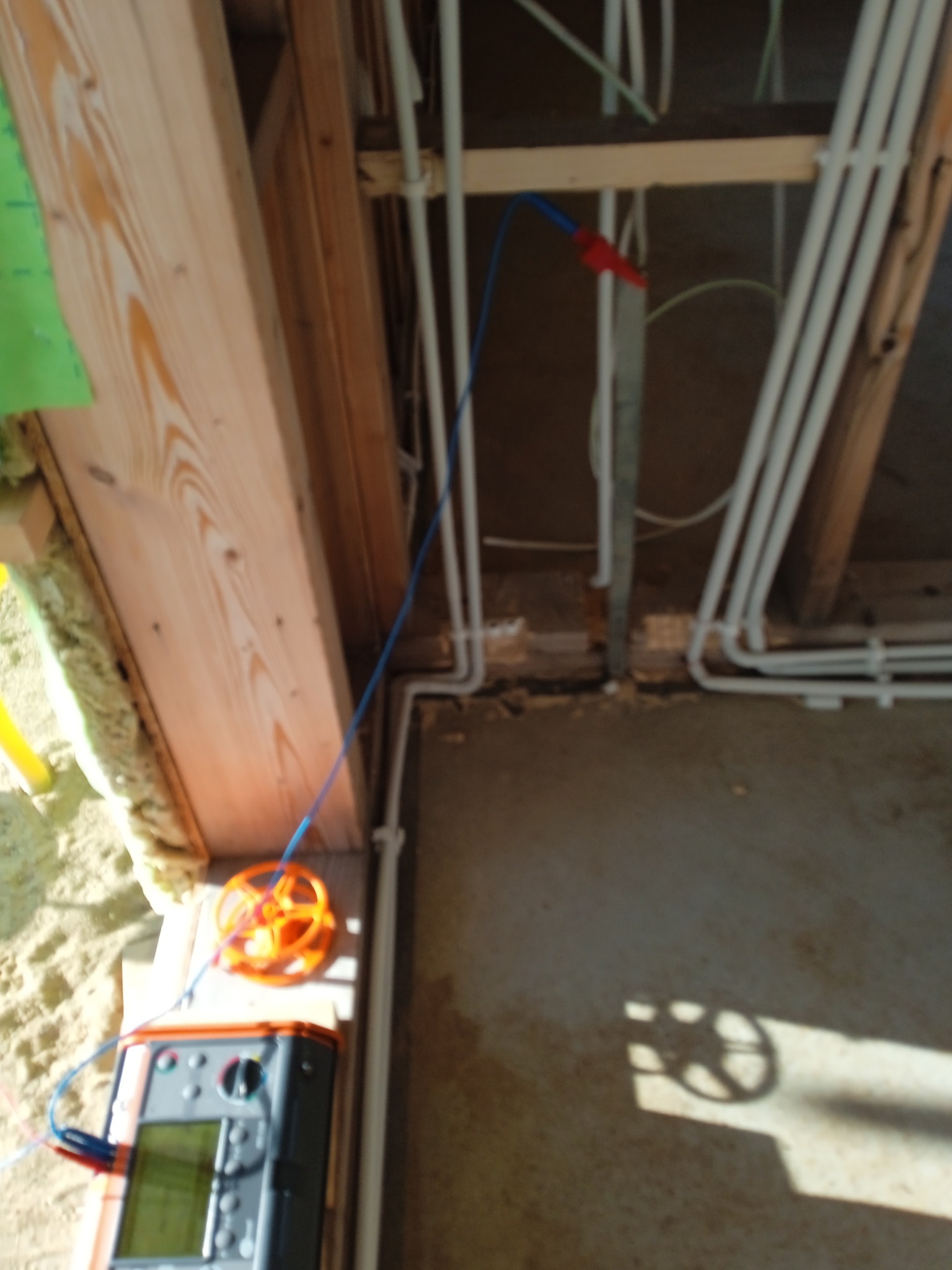 Pomiary instalacji elektrycznej w surowym wnętrzu. Widoczne przewody, miernik i elementy konstrukcji drewnianej. Niebieski przewód pomiarowy.