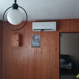 LifeTech Marcin Stawinoga - Biały klimatyzator Haier zamontowany na drewnianej ścianie w pomieszczeniu. Widoczny fragment lampy i mebli kuchennych. Ustawiona temperatura to 21 stopni.