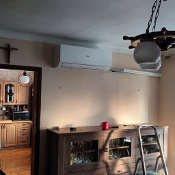 LifeTech Marcin Stawinoga - Zamontowany klimatyzator Haier Revive w jasnym wnętrzu. Widoczna szafka, drabina i mężczyzna przy otwartym balkonie. Uwagę zwraca lampa w stylu marynistycznym.