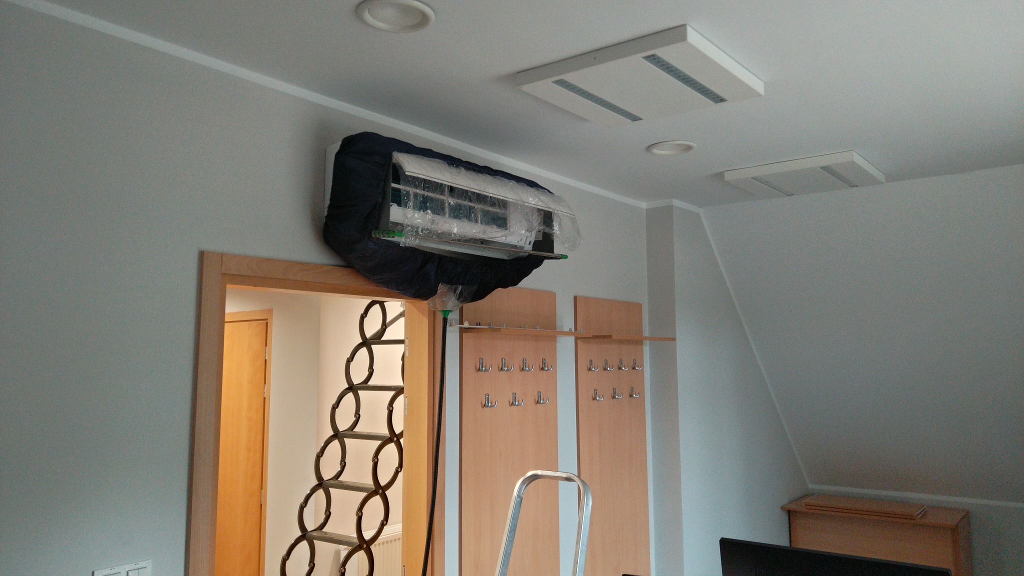 Klimatyzator zabezpieczony folią i pokrowcem podczas czyszczenia. Widoczna drabina i elementy garderoby w tle. Usługa dezynfekcji klimatyzacji.