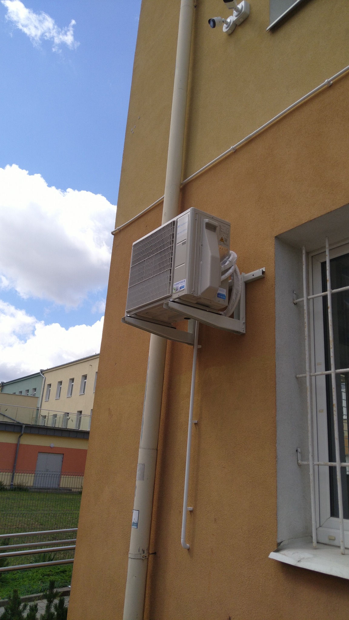 Zamontowany klimatyzator Fuji na elewacji budynku, widoczne rury odprowadzające. Obok okno z kratami, w tle budynek z zielenią. Ujęcie z dołu, błękitne niebo.