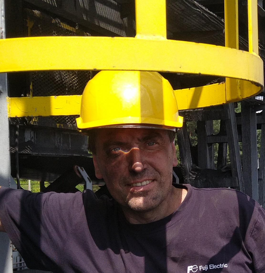 Uśmiechnięty instalator w kopalni węgla, w żółtym kasku, z zabrudzoną twarzą, po montażu klimatyzacji. Widoczne elementy konstrukcji kopalni.