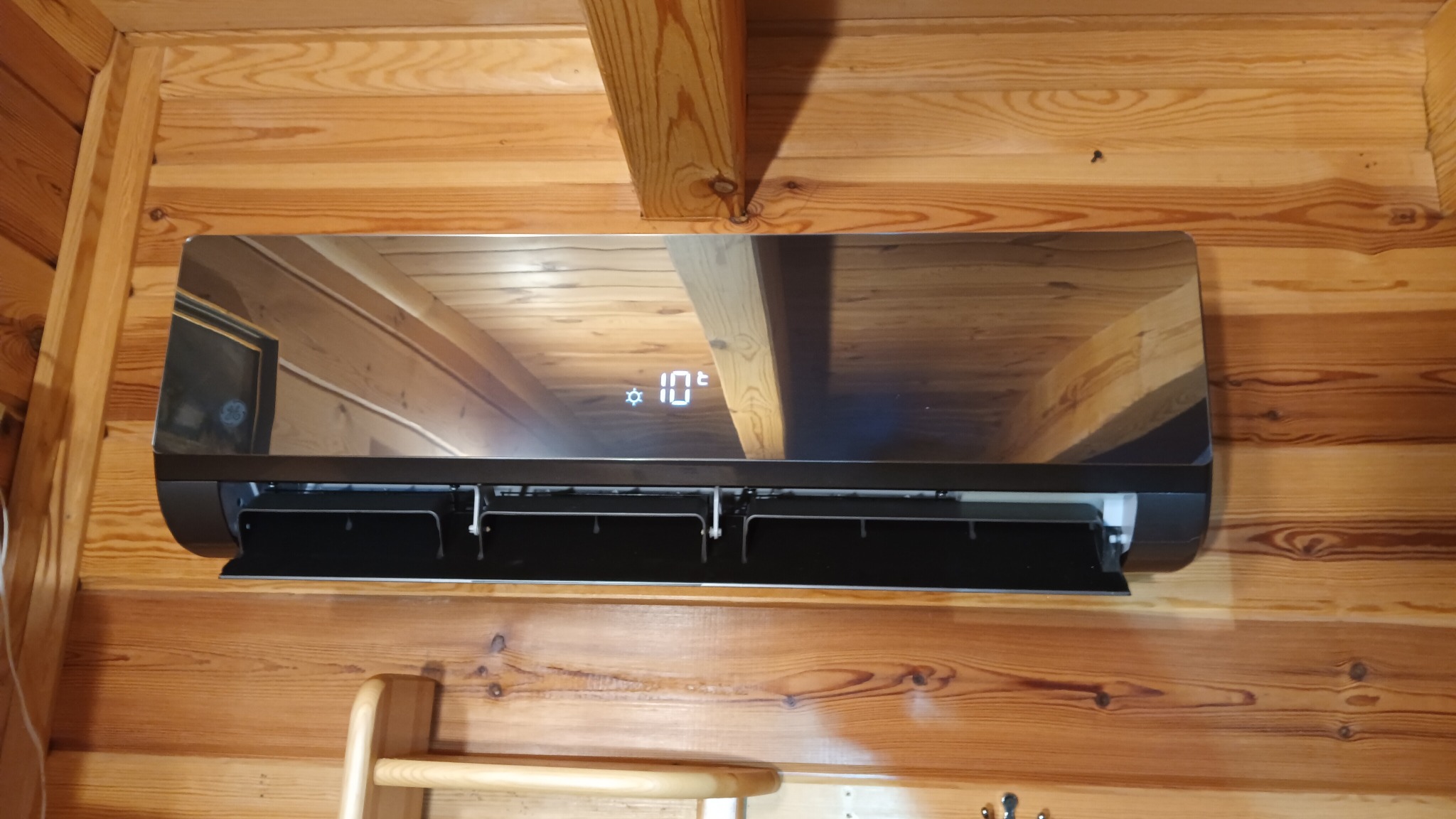 Zamontowany klimatyzator General Electric w drewnianym domku, widoczny wyświetlacz z temperaturą 10 stopni, odbicie drewnianej ściany w obudowie.