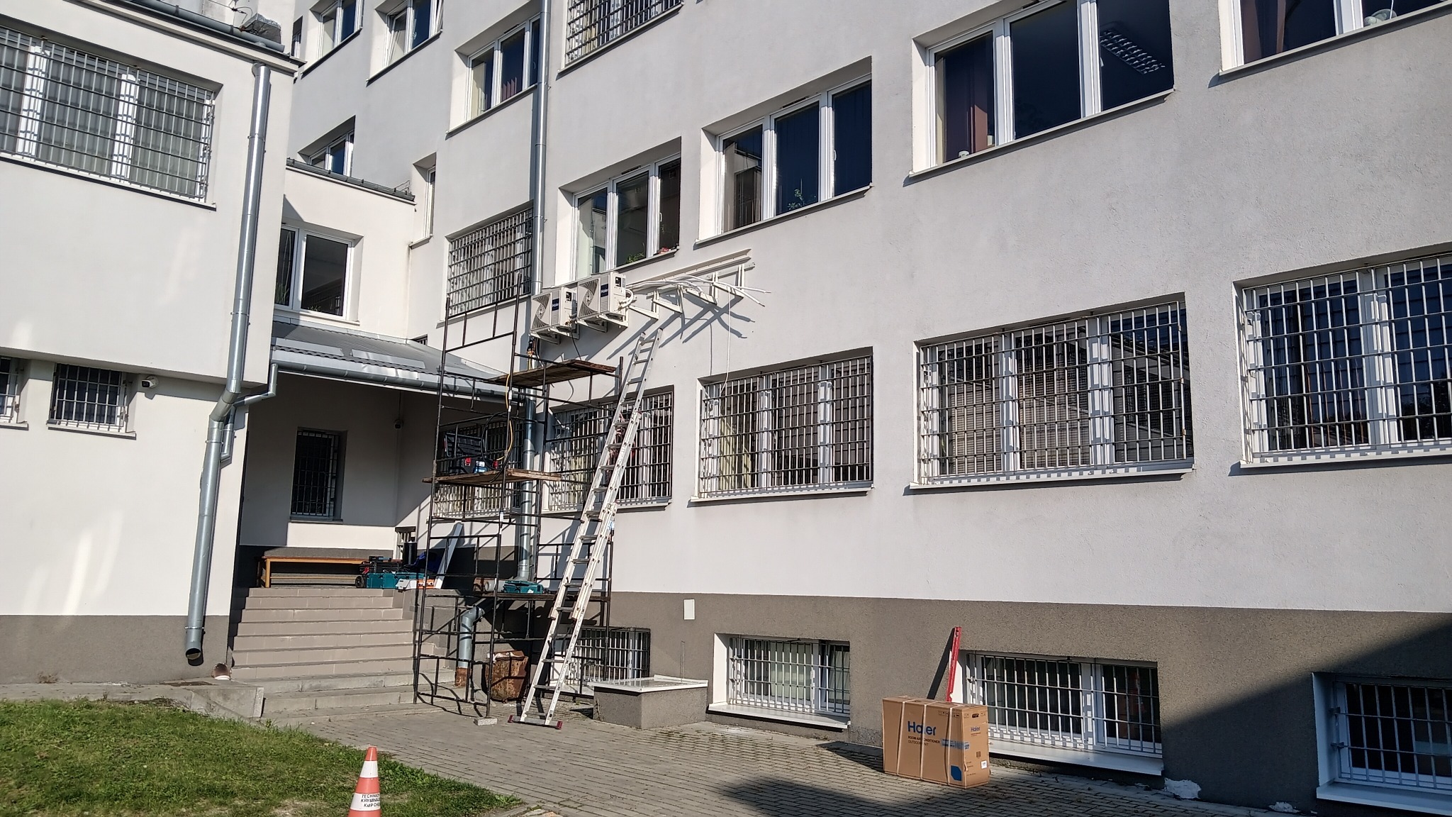 Montaż klimatyzatora Haier na elewacji budynku z oknami z kratami, widoczne rusztowanie i drabina. Karton po klimatyzatorze na chodniku. Słoneczny dzień.