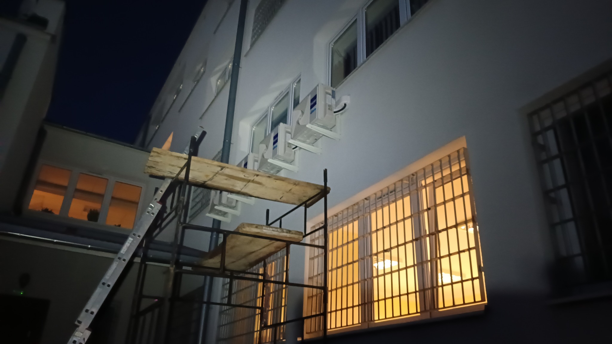 Montaż klimatyzatorów Haier na elewacji budynku z rusztowaniem w tle, widoczne okna z kratami, nocne oświetlenie.