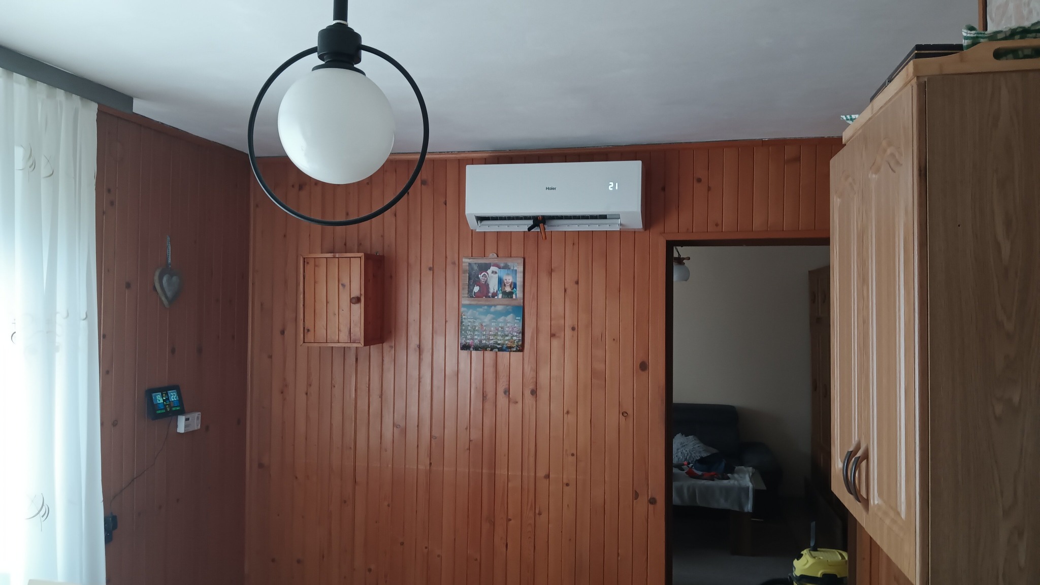 Biały klimatyzator Haier zamontowany na drewnianej ścianie w pomieszczeniu. Widoczny fragment lampy i mebli kuchennych. Ustawiona temperatura to 21 stopni.