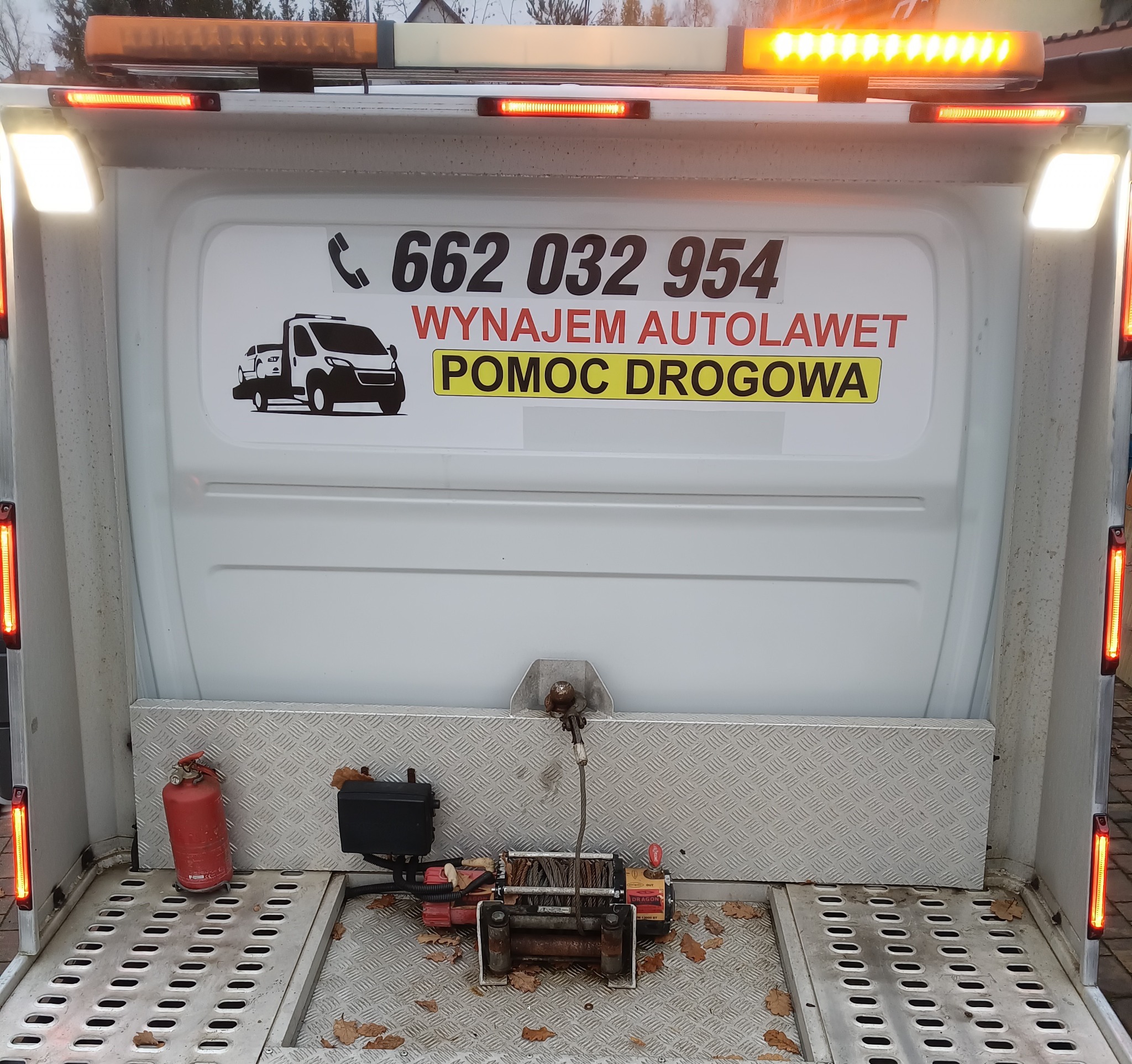 Tył lawety z wyciągarką, gaśnicą i naklejką z numerem telefonu oraz napisem 'Wynajem autolawet, Pomoc Drogowa'. Widoczne światła ostrzegawcze.