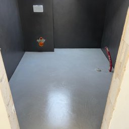 Pracownia Usługowa Agata Binkowska - Szara, gładka posadzka w łazience z ciemnymi ścianami, widok z progu. Instalacja wodna na ścianie. Skosy sufitowe. Minimalistyczne wykończenie.
