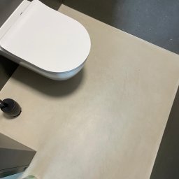 Pracownia Usługowa Agata Binkowska - Beżowa posadzka mikrocementowa w łazience z toaletą podwieszaną i szarą ścianą. Widok z góry.