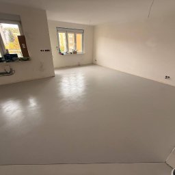 Pracownia Usługowa Agata Binkowska - Jasnoszara, gładka posadzka w pustym pokoju z dwoma oknami. Świeżo wylana, jednolita powierzchnia z widocznymi śladami po narzędziach. Przygotowanie do dalszych prac.