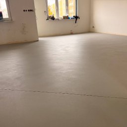 Pracownia Usługowa Agata Binkowska - Jasnoszara, gładka posadzka w surowym wnętrzu z oknami. Widoczne ślady po pracach wykończeniowych na ścianach. Minimalistyczny styl, świeżo wylana podłoga.