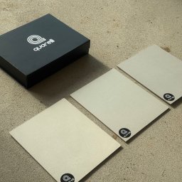 Pracownia Usługowa Agata Binkowska - Prezentacja próbek materiału wykończeniowego z logo producenta, ułożonych na surowym podłożu obok czarnego pudełka z logo. Neutralna kolorystyka, bliski kadr.