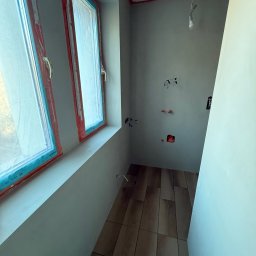 Pracownia Usługowa Agata Binkowska - Wnętrze w trakcie prac wykończeniowych w Trzebnicy. Widoczne okna z folią ochronną, położona podłoga, ściany przygotowane do malowania, instalacja elektryczna.