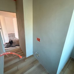 Pracownia Usługowa Agata Binkowska - Ściana po nałożeniu mikrocementu w odcieniach szarości, widoczne instalacje elektryczne i hydrauliczne, podłoga wyłożona panelami. W tle pomieszczenie zabezpieczone folią.