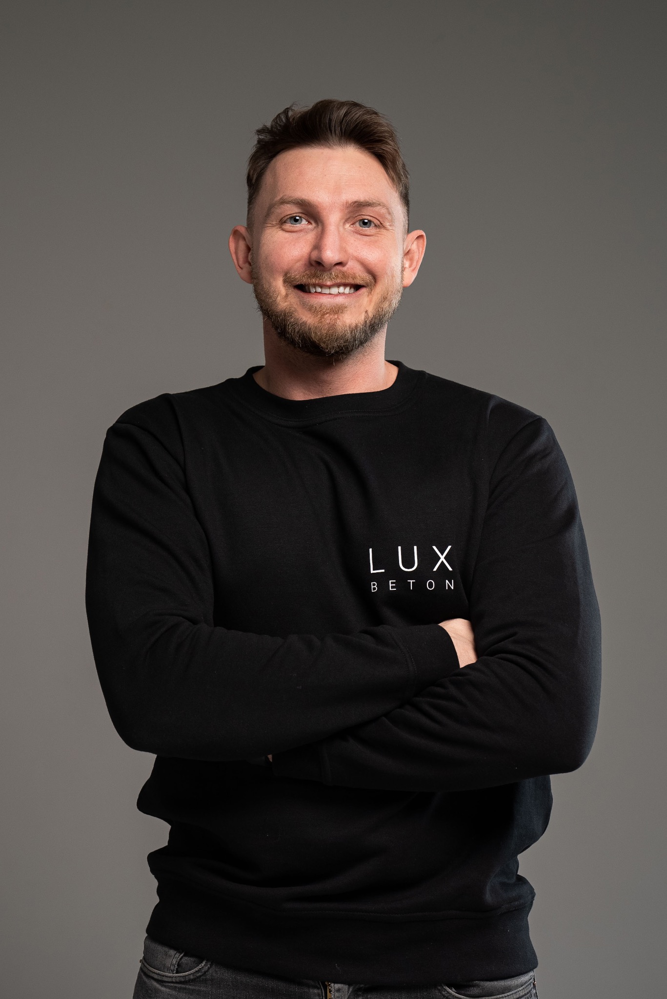 Portret uśmiechniętego mężczyzny w czarnej bluzie z logo firmy z Trzebnicy, na szarym tle. Mężczyzna ma brodę i niebieskie oczy.