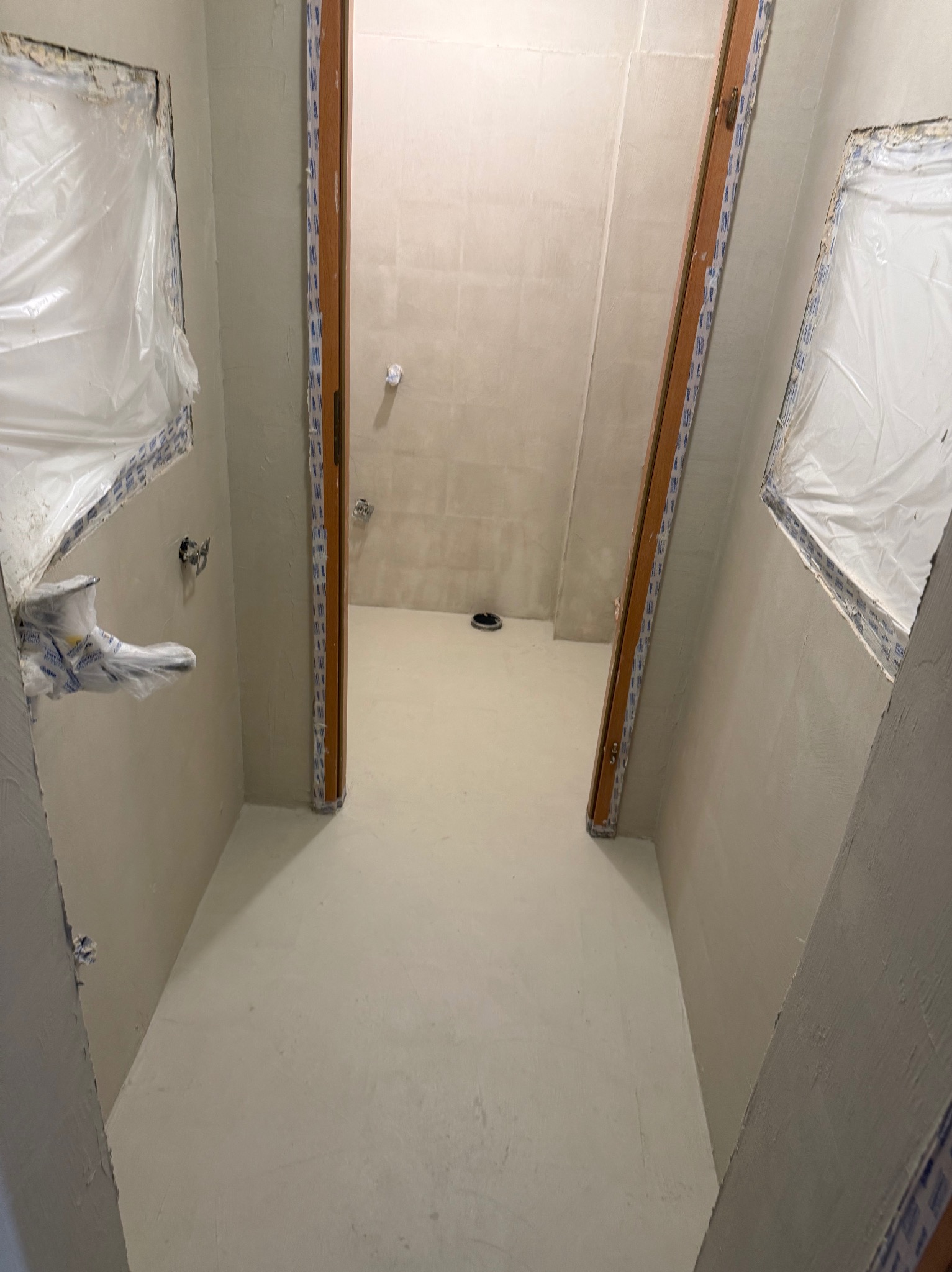 Wnętrze łazienki w trakcie prac wykończeniowych: zabezpieczone okna folią, świeżo położona jasna wylewka, przygotowane ściany pod dalsze prace. Widok z perspektywy wejścia.