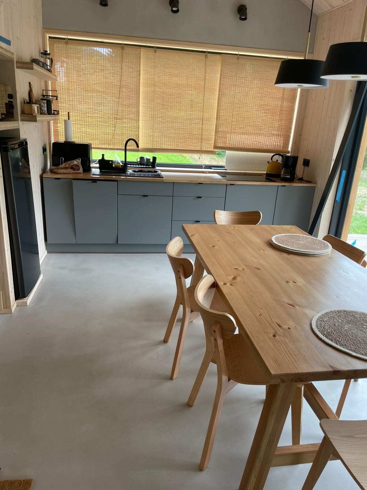 Minimalistyczna kuchnia z drewnianym stołem i szarą posadzką w Trzebnicy. Widoczne rolety bambusowe i nowoczesne oświetlenie.