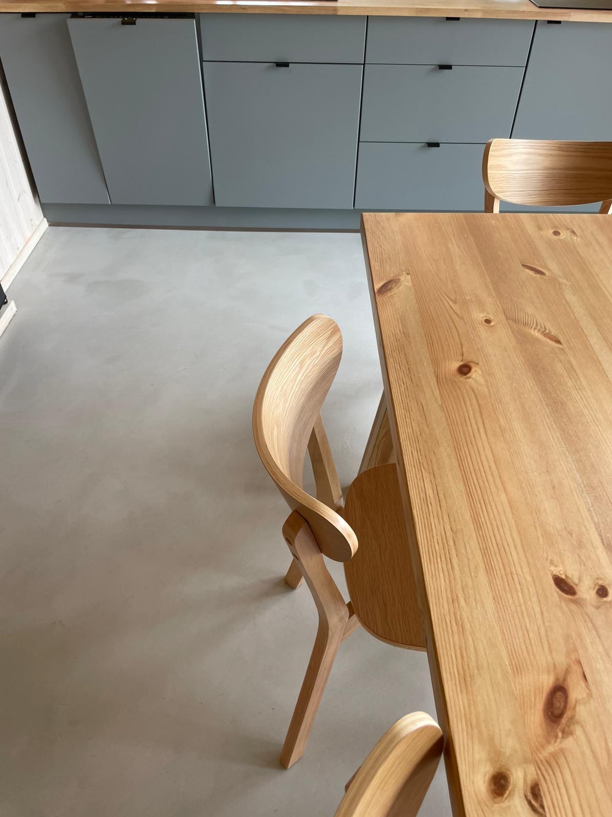 Jasnoszara posadzka w minimalistycznej kuchni z drewnianym stołem i krzesłami. Blaty kuchenne w tle o jednolitej kolorystyce.