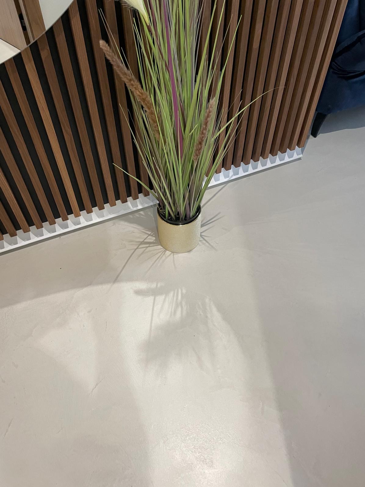 Jasna, jednolita posadzka w neutralnym odcieniu, z dekoracyjną rośliną w złotej doniczce. W tle drewniane panele ścienne z czarnymi wstawkami.