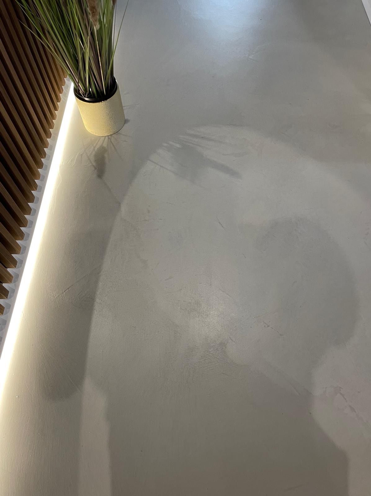 Jasnoszara posadzka mikrocementowa z subtelną teksturą, oświetlona listwą LED przy drewnianej ściance. W rogu doniczka z trawą, rzucająca cień na gładką powierzchnię.