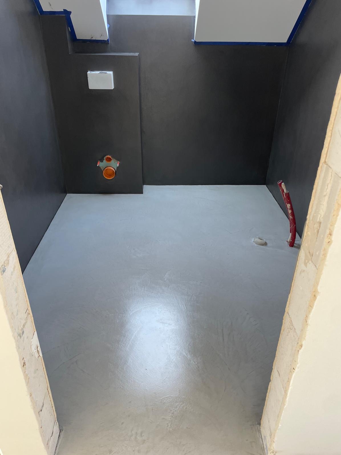 Szara, gładka posadzka w łazience z ciemnymi ścianami, widok z progu. Instalacja wodna na ścianie. Skosy sufitowe. Minimalistyczne wykończenie.