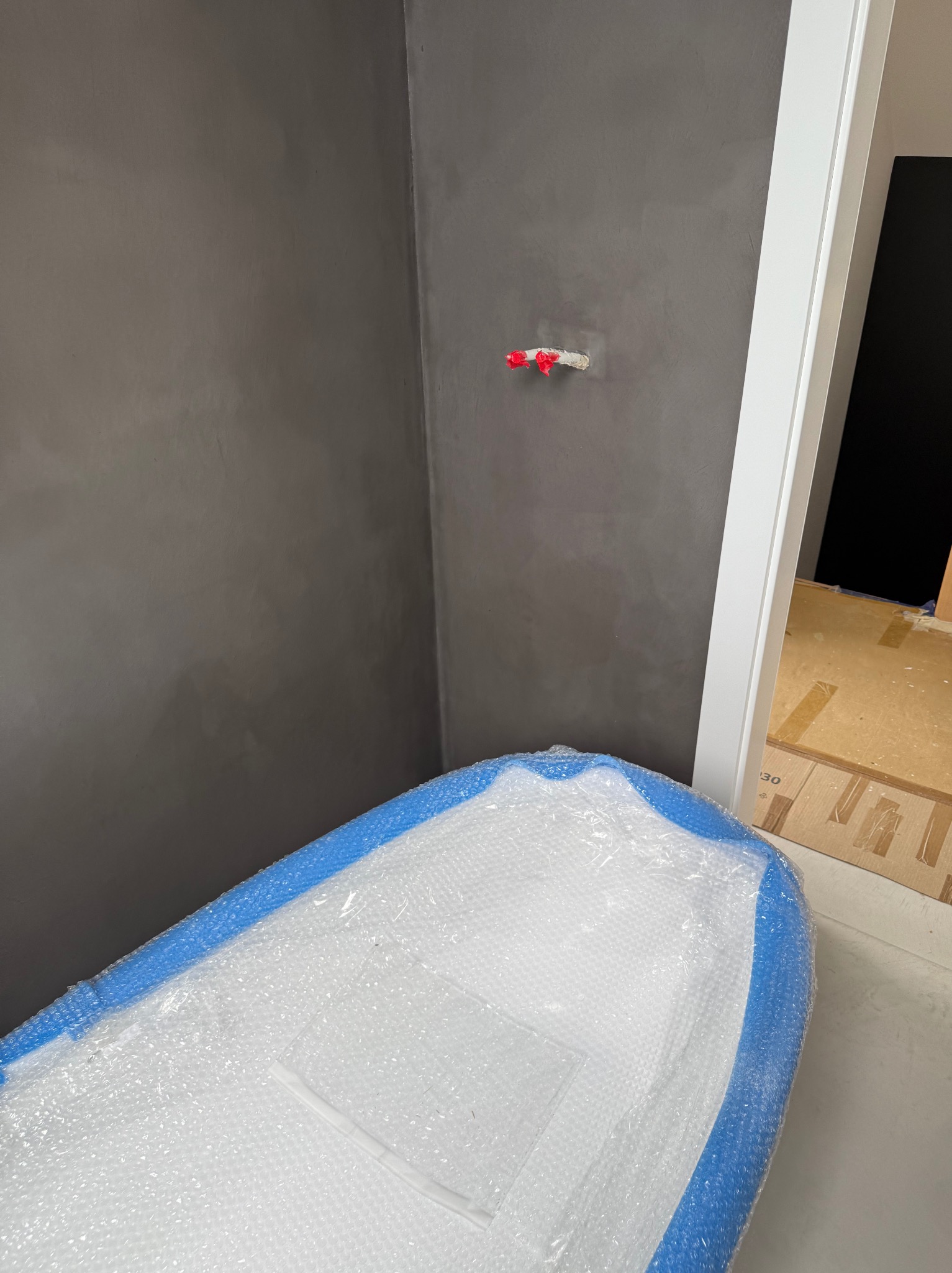 Szara ściana z gładzią i zabezpieczona folią wanna z błękitnym obramowaniem, fragment białej futryny drzwiowej. Widoczny wystający zawór.