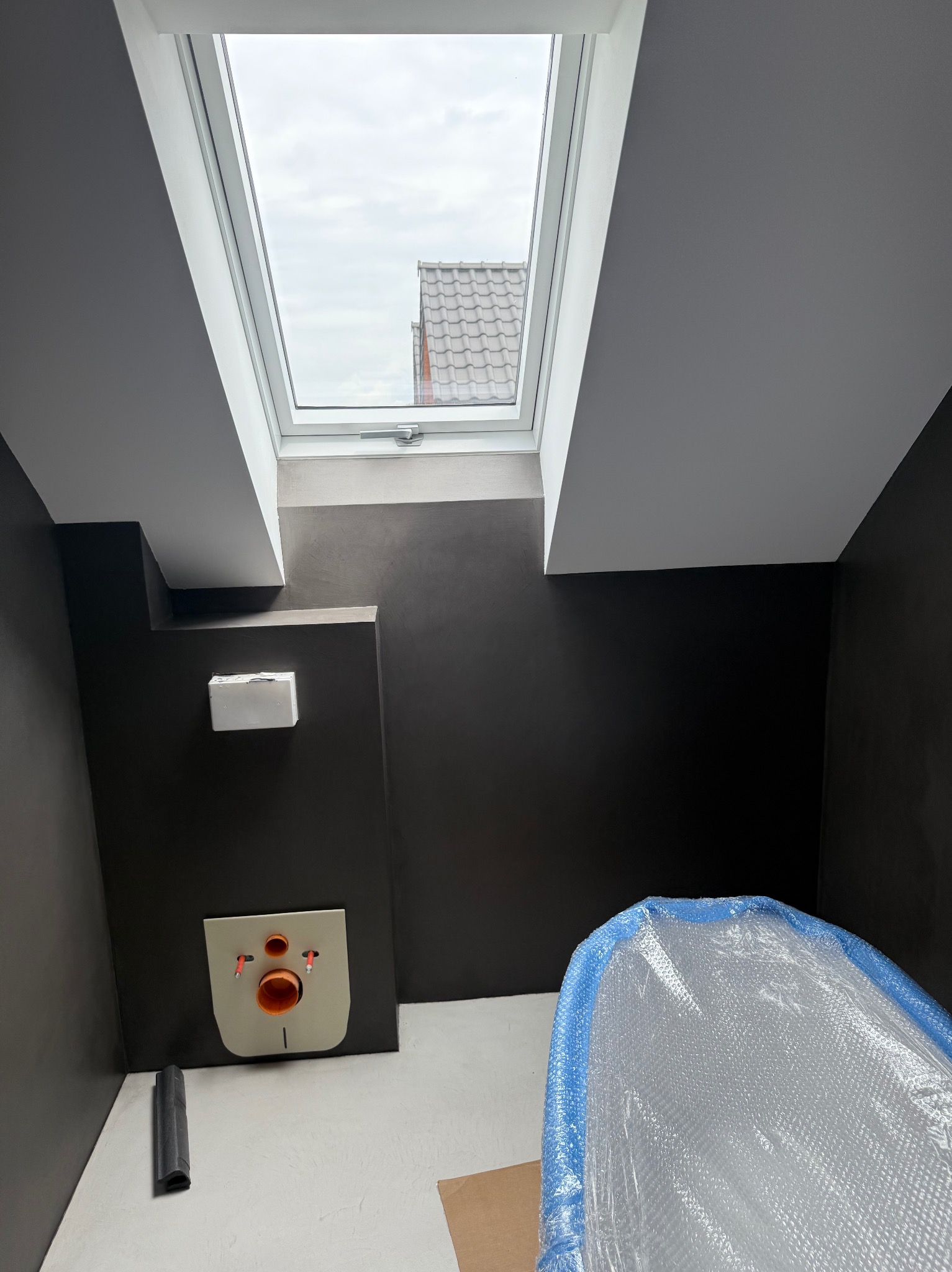 Wykończona łazienka na poddaszu z oknem dachowym. Ciemne ściany, biała podłoga, zabudowa WC i wanna zabezpieczona folią bąbelkową. Minimalistyczny design.
