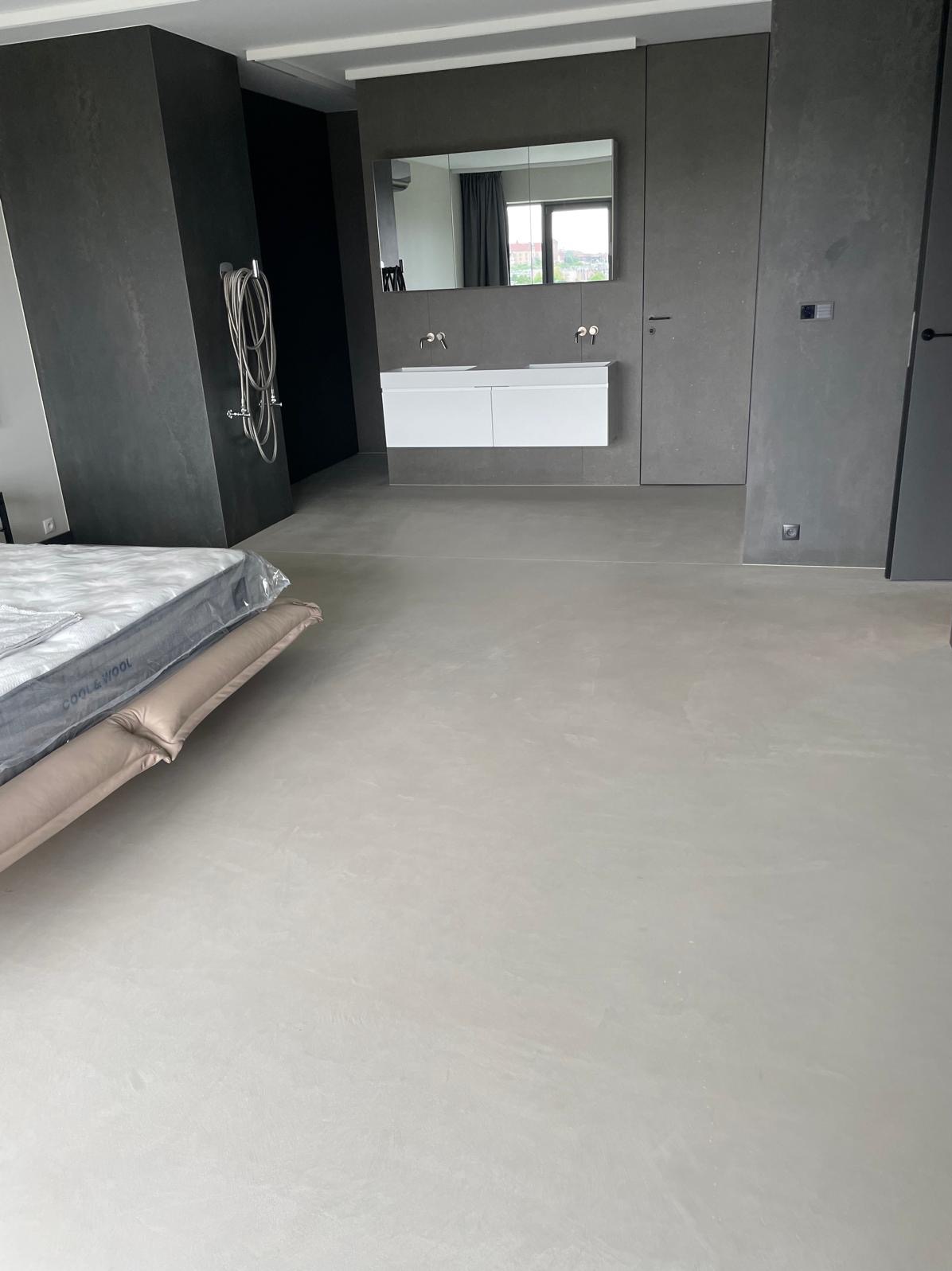 Minimalistyczna łazienka z szarą, jednolitą posadzką i ścianami w odcieniach szarości. Biała szafka z umywalkami pod lustrem. Widoczny fragment materaca.