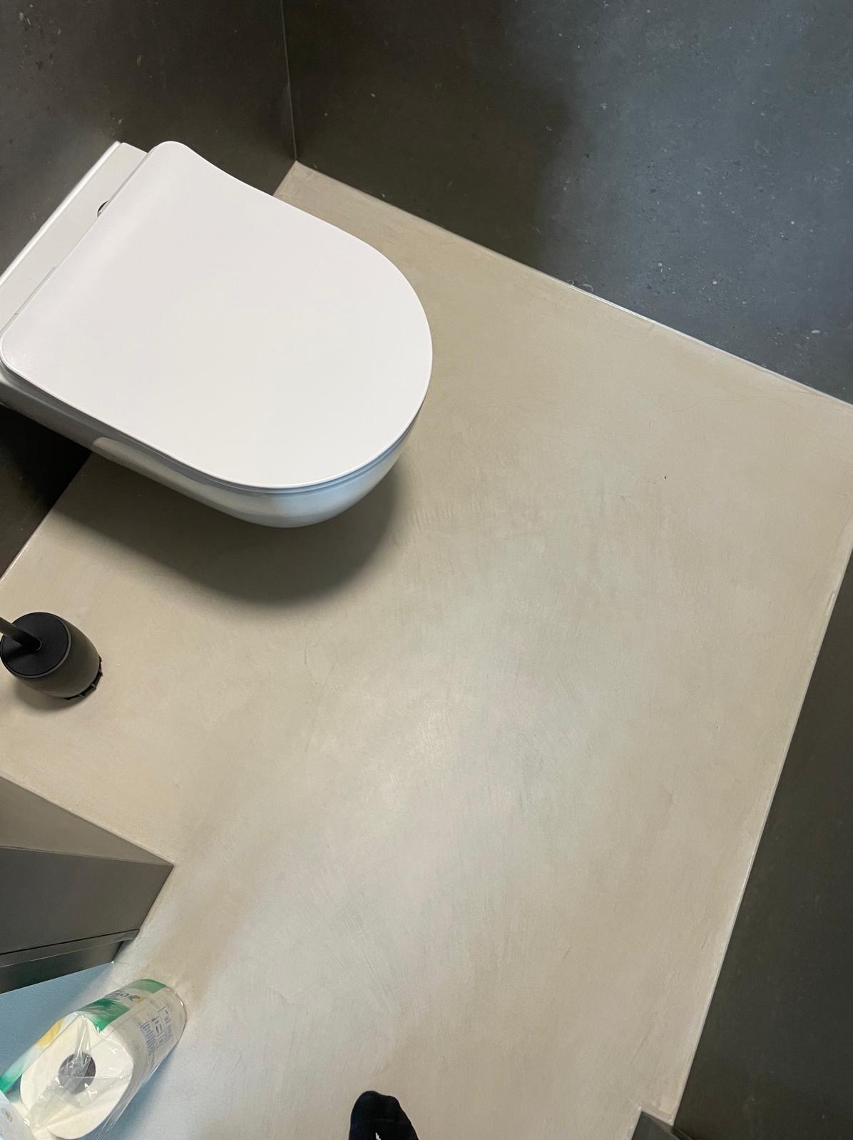 Beżowa posadzka mikrocementowa w łazience z toaletą podwieszaną i szarą ścianą. Widok z góry.