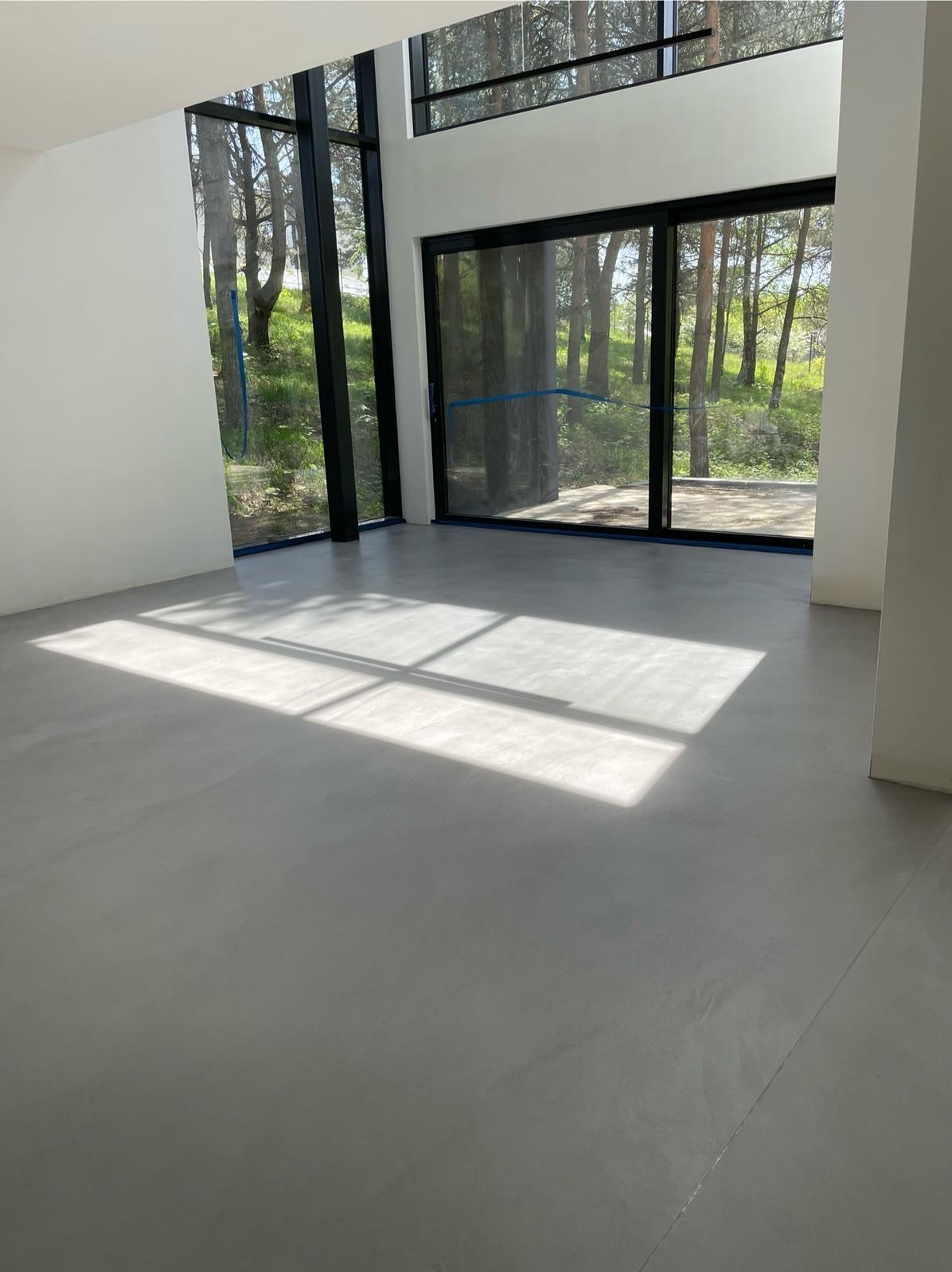 Jasnoszara, gładka podłoga w nowoczesnym salonie z dużymi oknami na las. Światło słoneczne tworzy geometryczne wzory na posadzce.