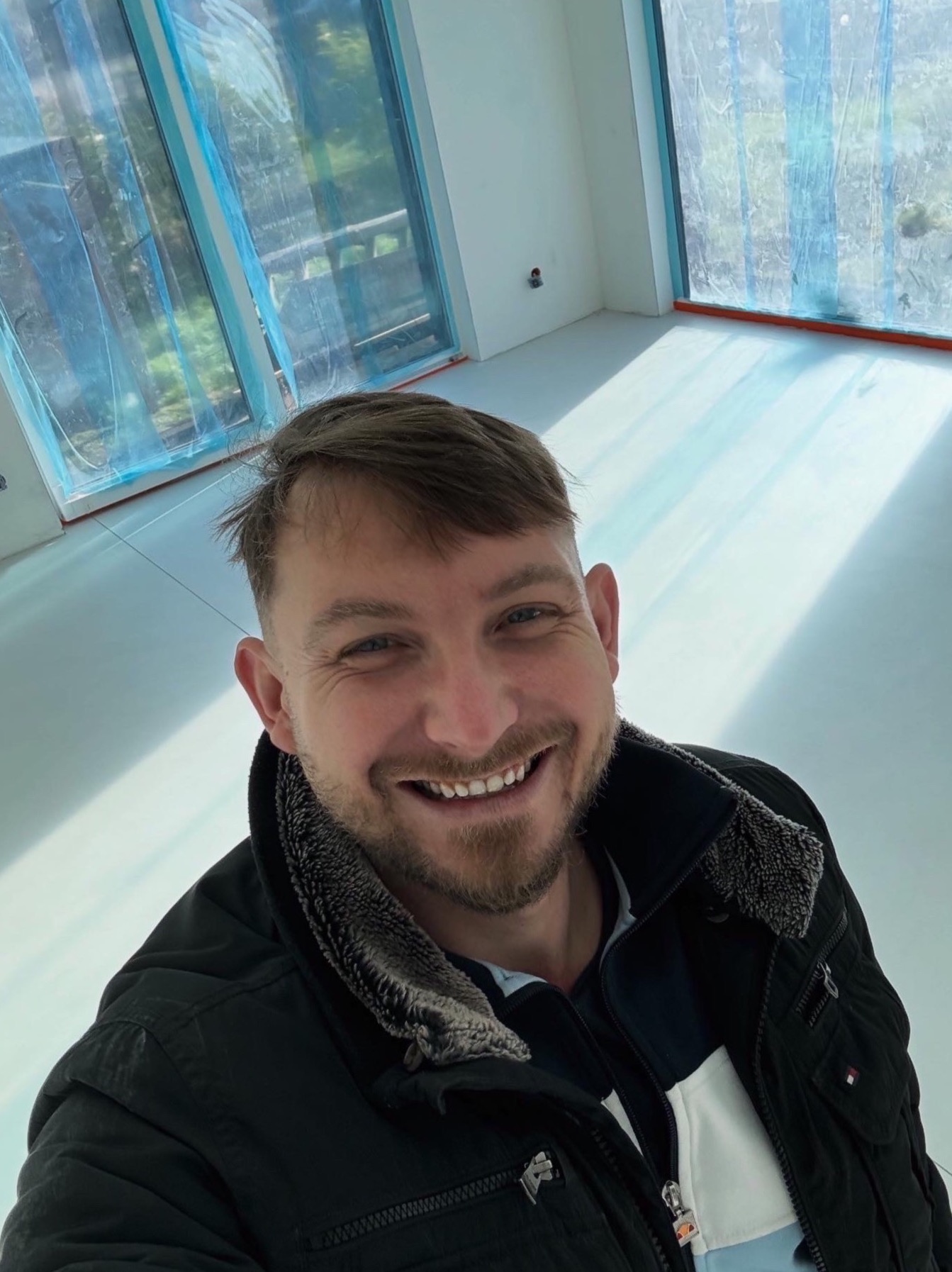 Uśmiechnięty mężczyzna na tle jasnej, gładkiej posadzki w nowym wnętrzu z dużymi oknami zabezpieczonymi folią. Widoczny fragment kurtki zimowej.