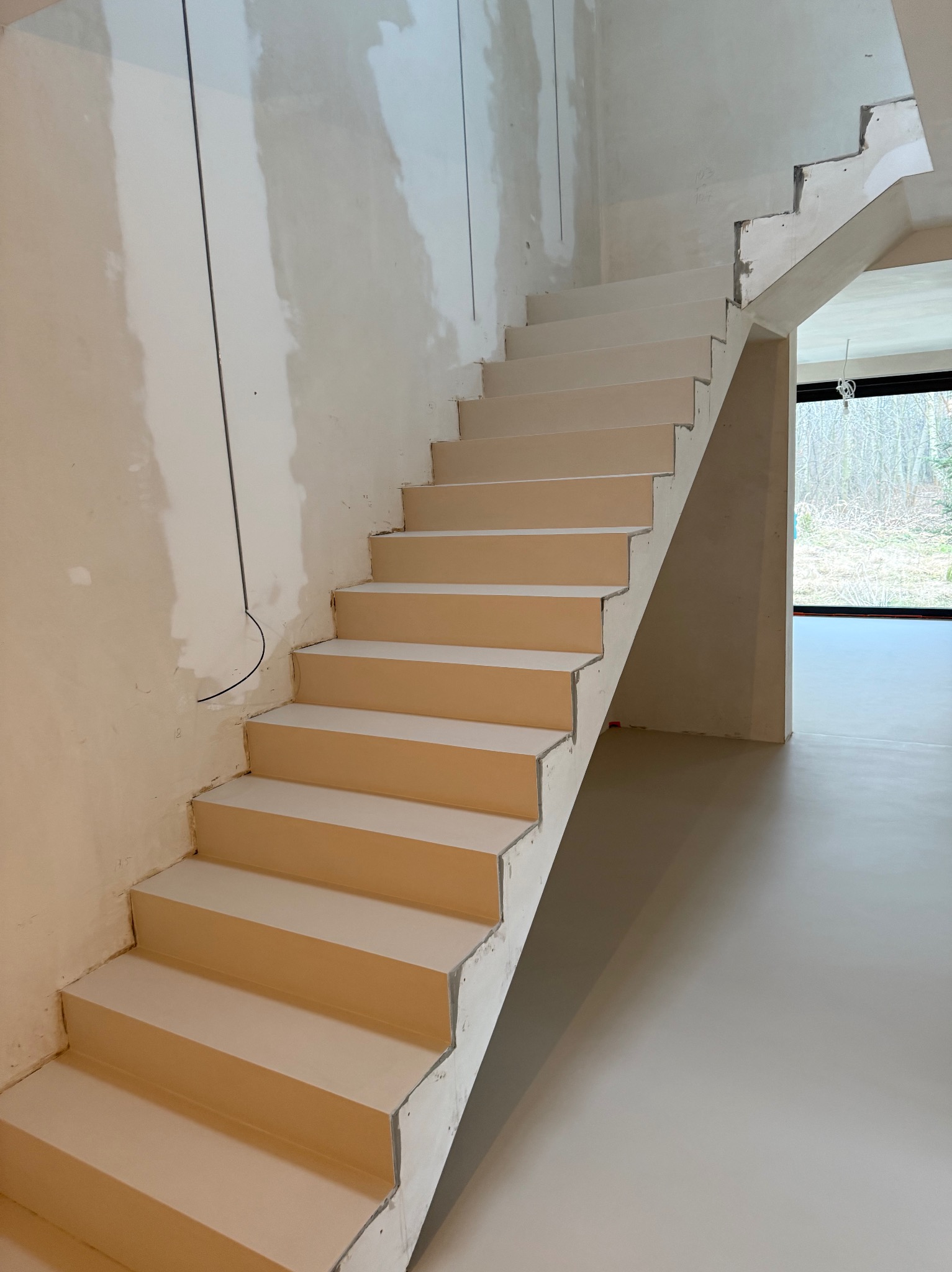 Jasne schody w surowym wnętrzu z widoczną wylewką podłogową i oknem. Widoczne niedoskonałości ścian i brak listew przypodłogowych. Minimalistyczny design w trakcie prac.