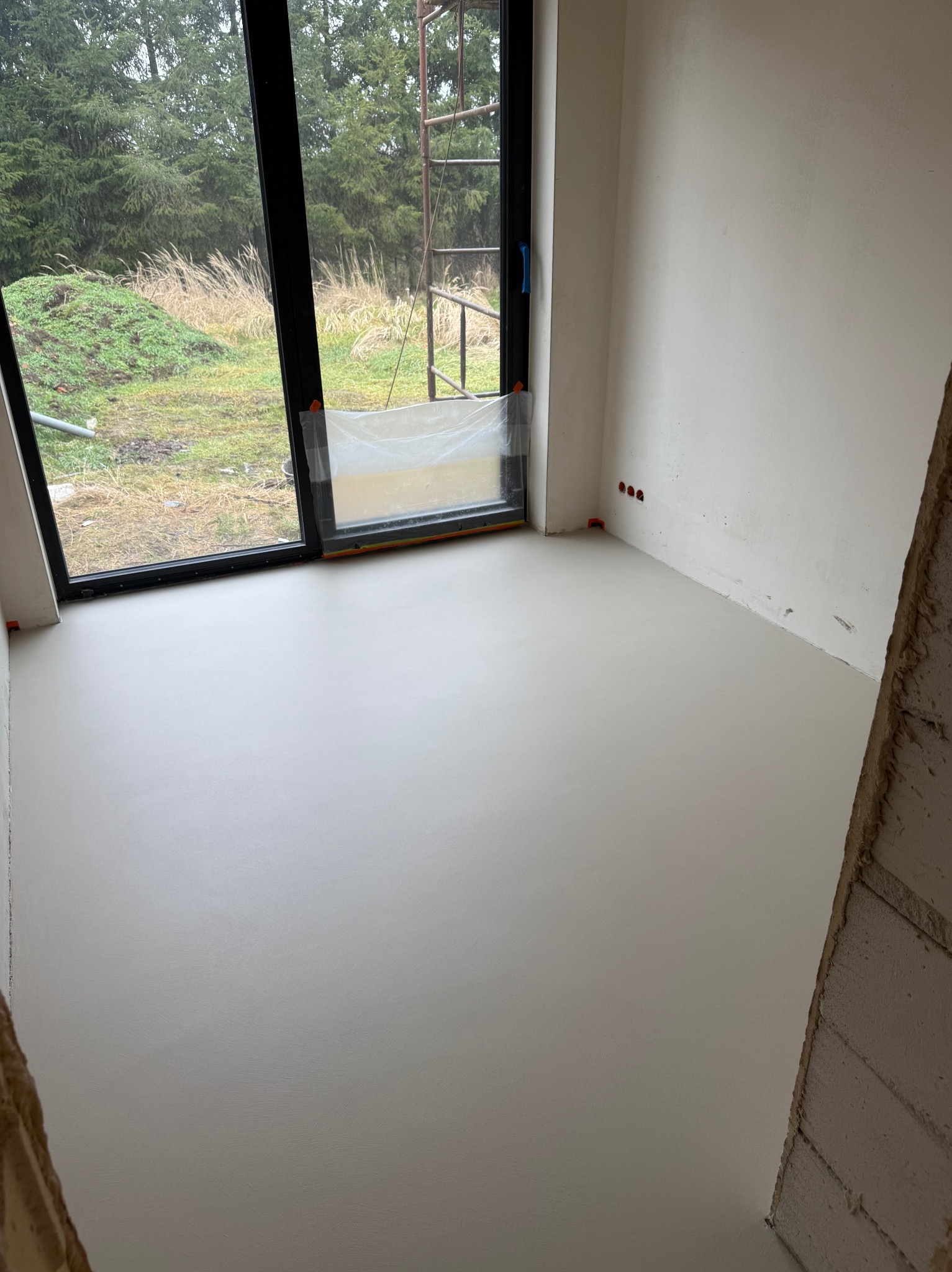 Jasnoszara, gładka posadzka w surowym wnętrzu z dużym oknem na zewnątrz. Widoczne ślady prac wykończeniowych na ścianach i narożnikach. Minimalistyczny styl.