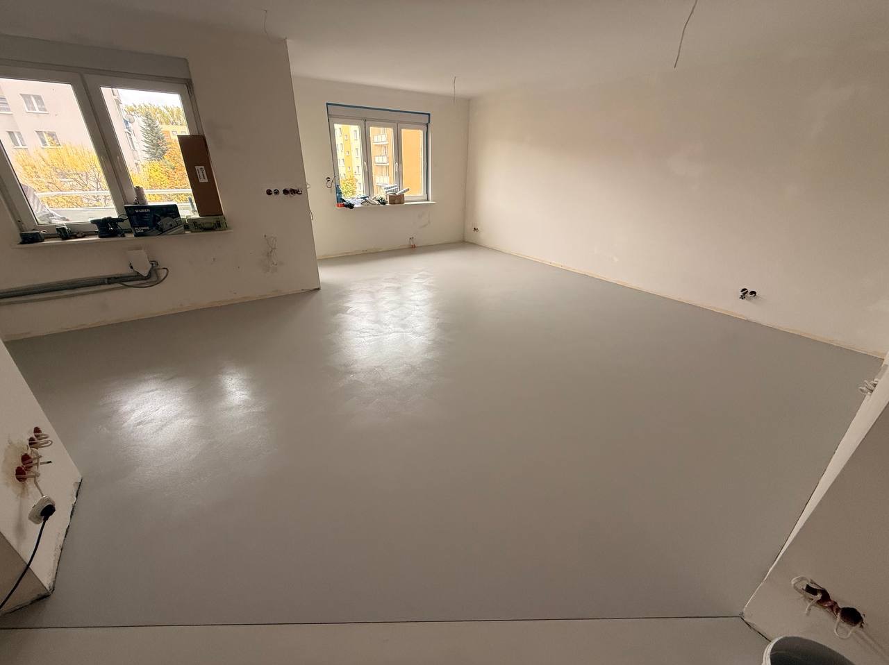 Jasnoszara, gładka posadzka w pustym pokoju z dwoma oknami. Świeżo wylana, jednolita powierzchnia z widocznymi śladami po narzędziach. Przygotowanie do dalszych prac.