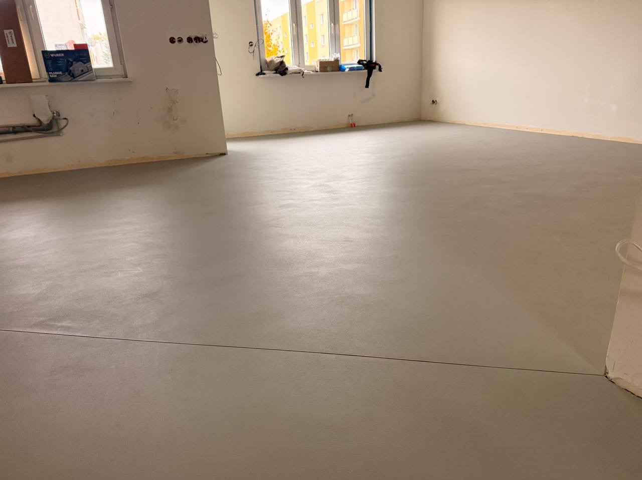 Jasnoszara, gładka posadzka w surowym wnętrzu z oknami. Widoczne ślady po pracach wykończeniowych na ścianach. Minimalistyczny styl, świeżo wylana podłoga.