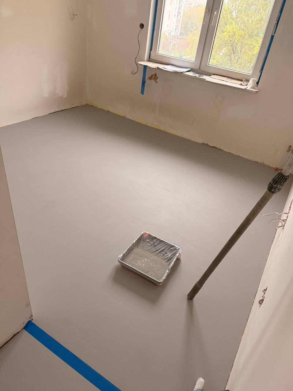 Świeżo wylana, gładka posadzka w jasnym odcieniu szarości w pokoju z oknem. Widoczna kuweta z resztkami materiału i wałek malarski. Niebieska taśma zabezpieczająca przy ścianie.