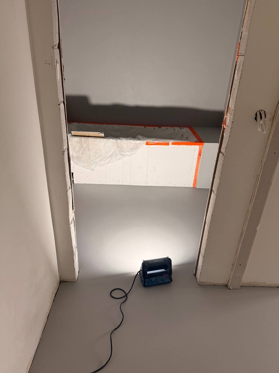 Szara, gładka posadzka w trakcie prac wykończeniowych, oświetlona lampą budowlaną. Widoczne przygotowania do dalszych prac na ścianach i suficie.