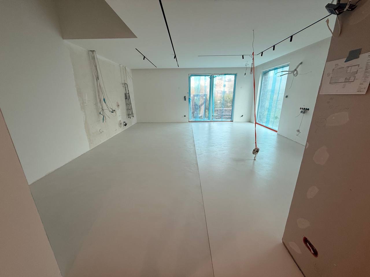 Jasna, gładka posadzka w przestronnym wnętrzu z widocznymi instalacjami na ścianach i oknami zabezpieczonymi folią. Minimalistyczne oświetlenie sufitowe.