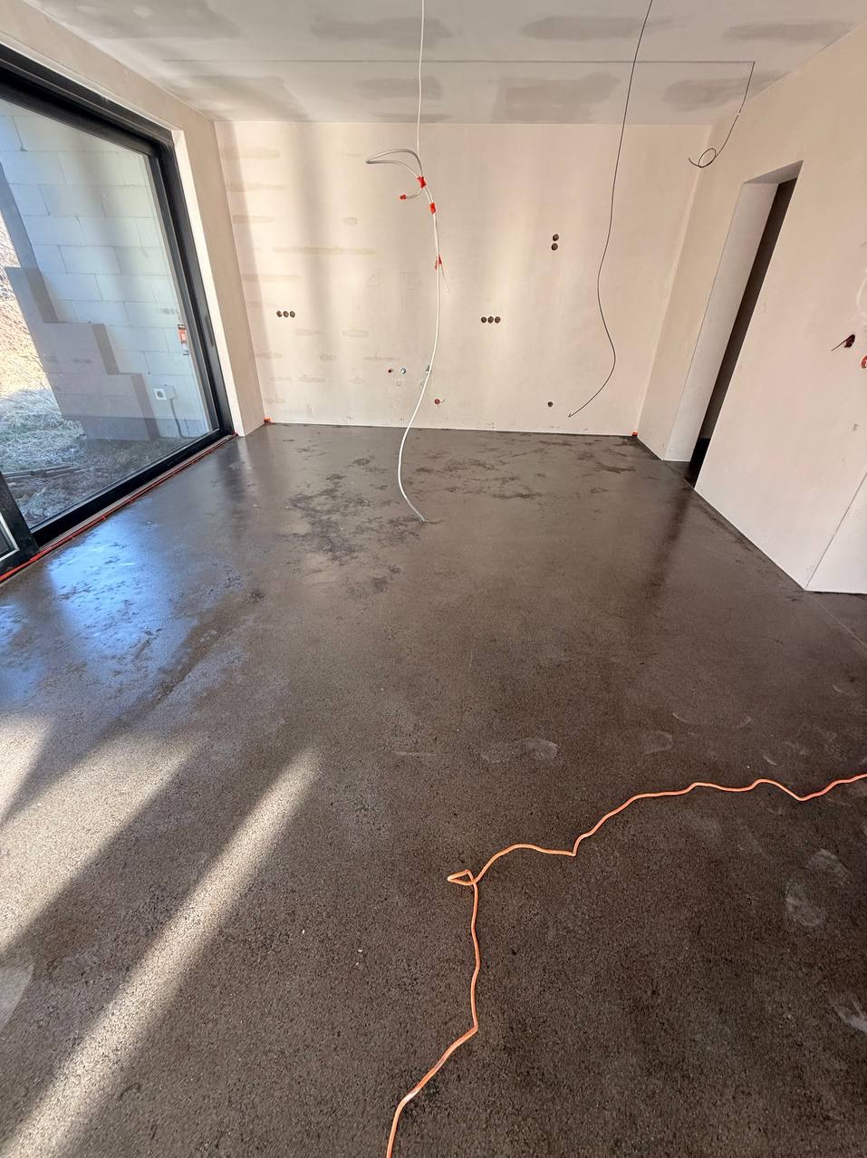 Szara, gładka posadzka w surowym wnętrzu z widocznymi przewodami elektrycznymi i dużym oknem. Świeżo wylana podłoga odbija światło, tworząc nowoczesny, minimalistyczny efekt.