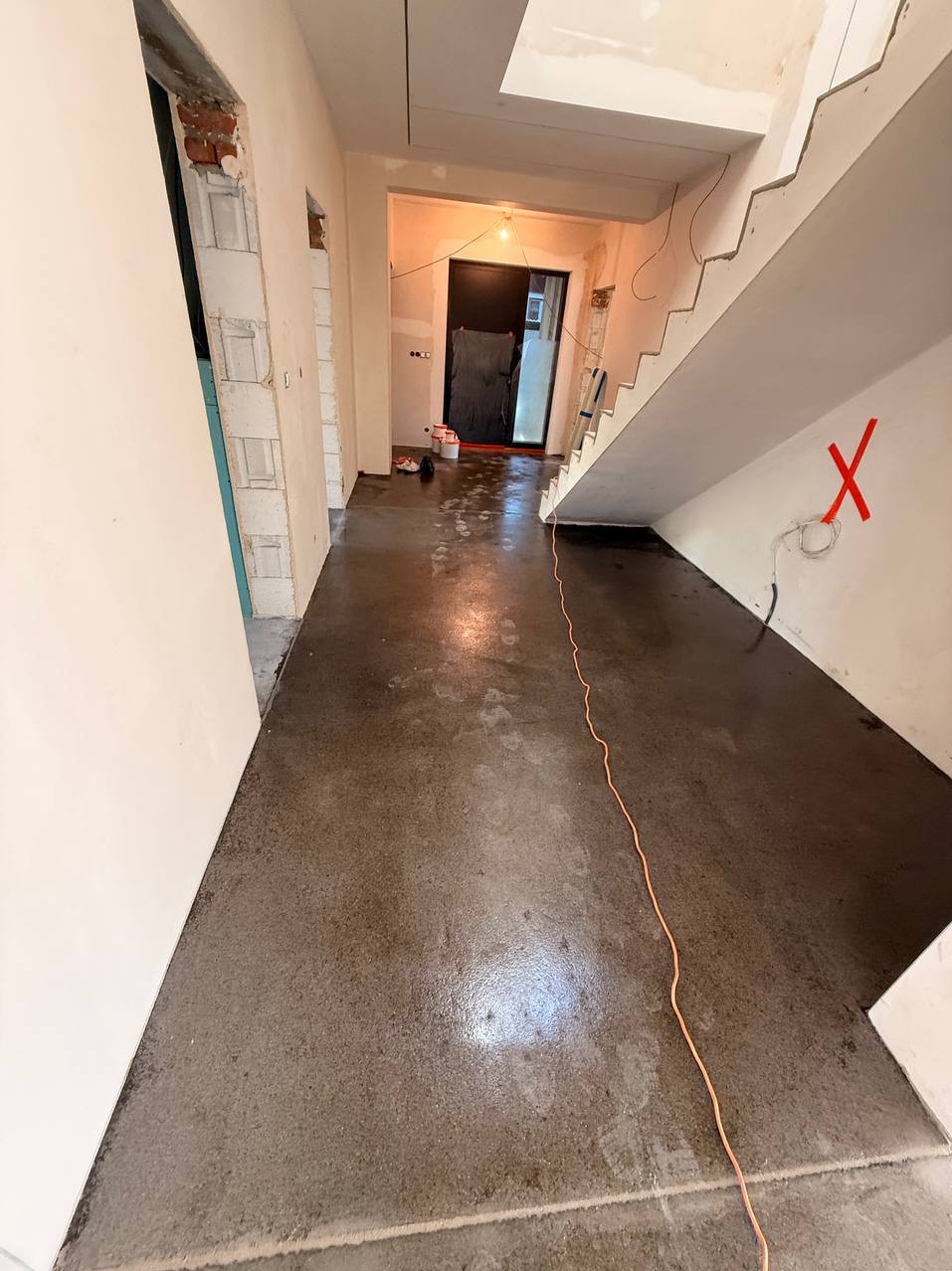 Błyszcząca, szara posadzka w surowym wnętrzu z widocznymi śladami stóp i kablem na podłodze. W tle schody i otwory drzwiowe w stanie deweloperskim.