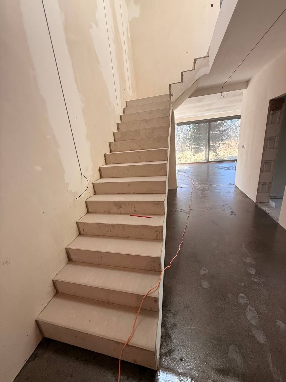 Surowe schody w jasnym wnętrzu w trakcie prac wykończeniowych. Widoczna wylewka na podłodze i ściany przygotowane do malowania. Duże okno w tle.