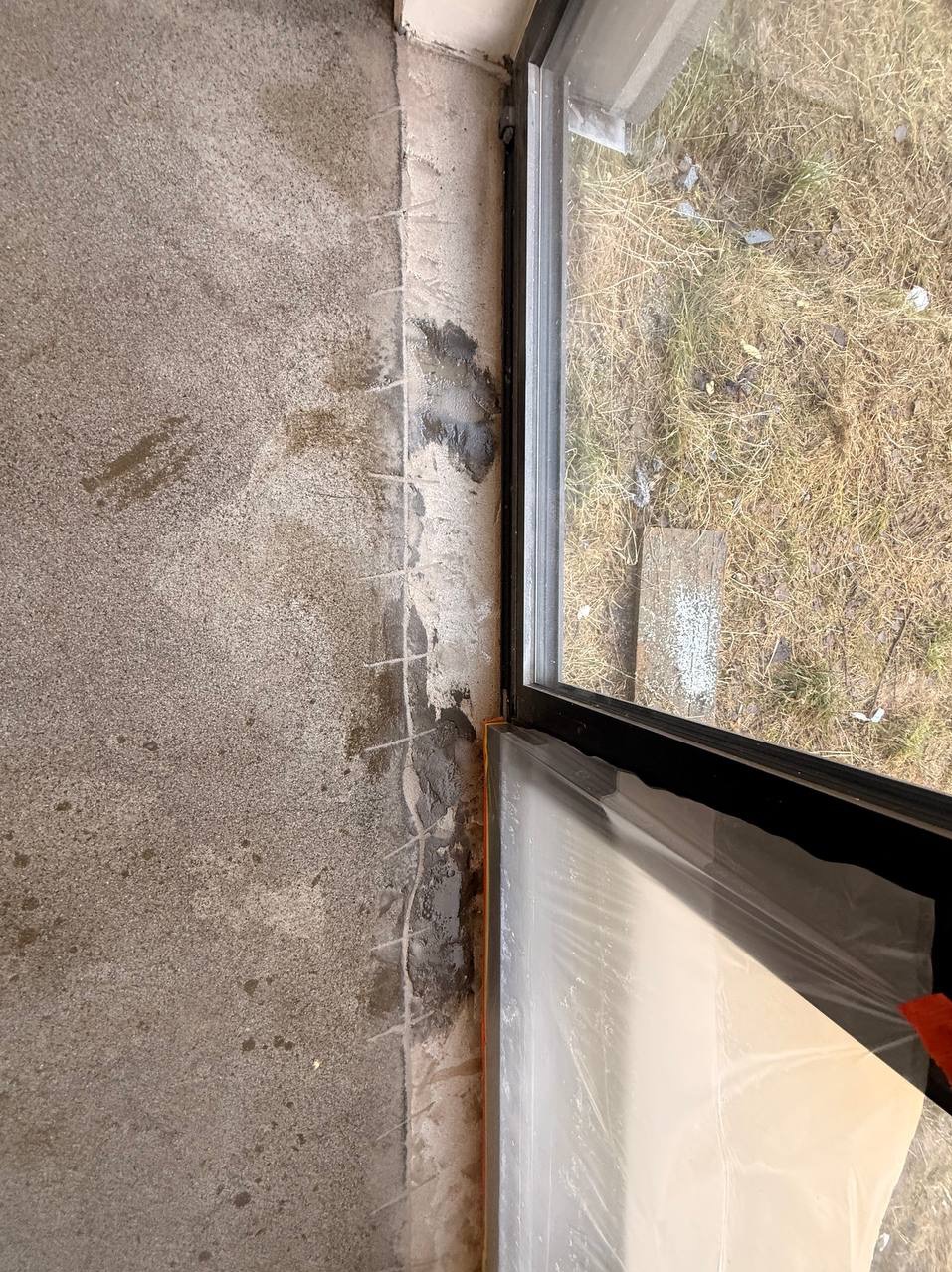 Szara, surowa posadzka przy oknie z widocznymi śladami po usuniętej listwie przypodłogowej i zabezpieczoną folią szybą, ukazująca etap prac wykończeniowych.