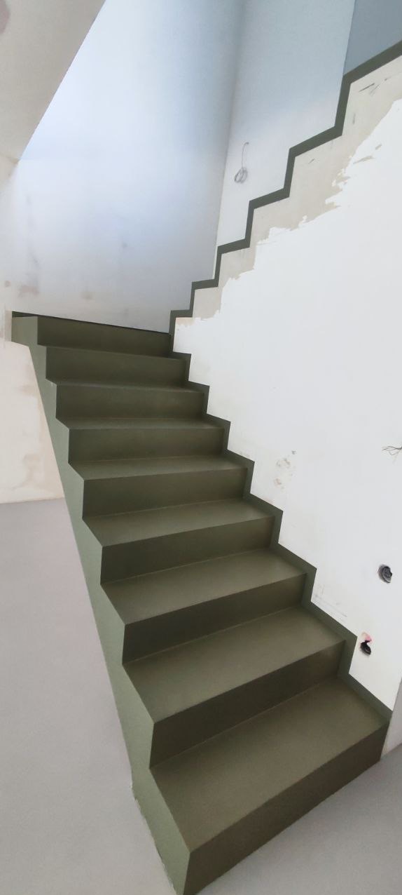 Schody wewnętrzne w odcieniach szarości, wykończone w minimalistycznym stylu. Widoczne ślady prac wykończeniowych na ścianie. Ujęcie z dołu, podkreślające geometrię.