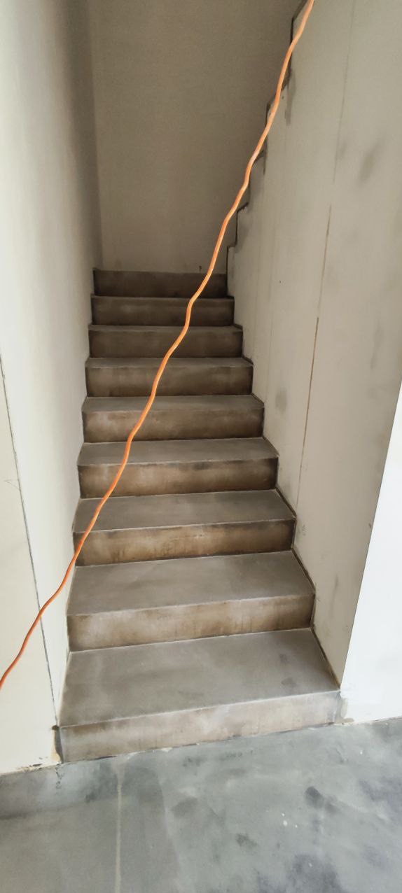 Betonowe schody w surowym stylu, z widocznym kablem elektrycznym. Minimalistyczne wykończenie wnętrza, szare odcienie, proste linie. Widok z dołu na górę.