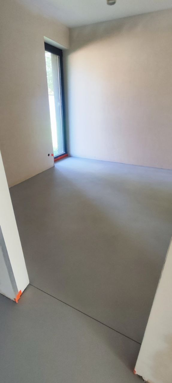 Szara, gładka posadzka w jasnym pokoju z oknem. Widoczne listwy przypodłogowe z pomarańczowymi zabezpieczeniami. Minimalistyczny design, świeżo wylana posadzka.