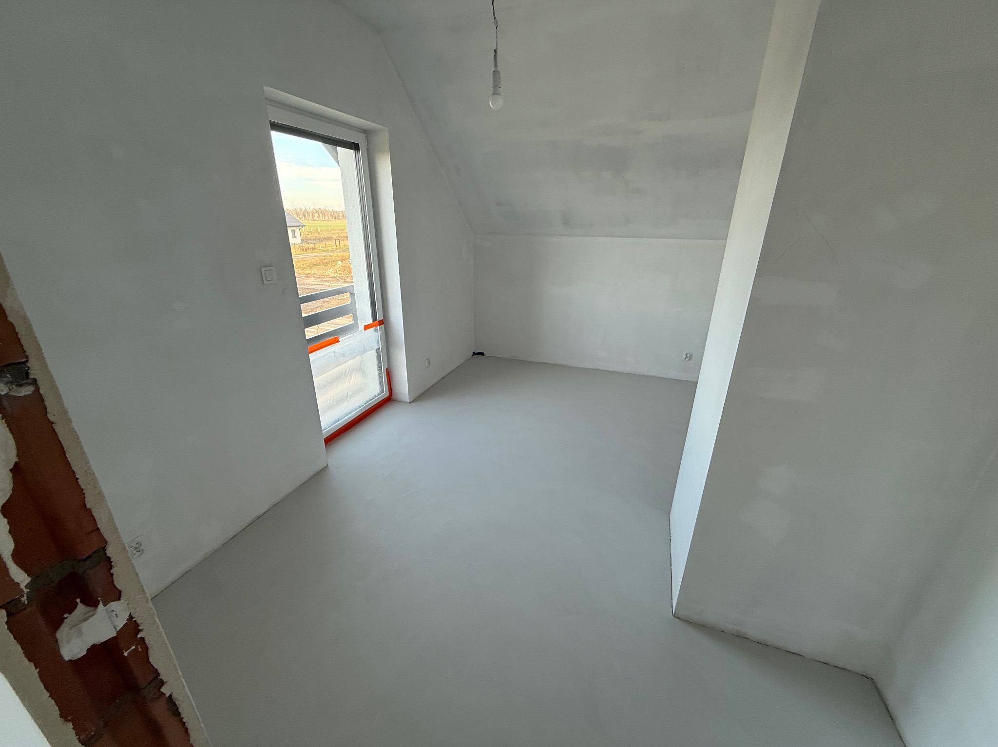 Jasne, surowe wnętrze poddasza z oknem i świeżo wylaną, gładką posadzką w odcieniach szarości. Widoczny fragment ściany z cegły w lewym dolnym rogu kadru.